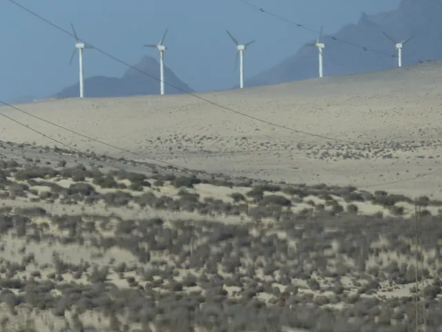 Wind turbines on pale dunes with jagged mountains behind