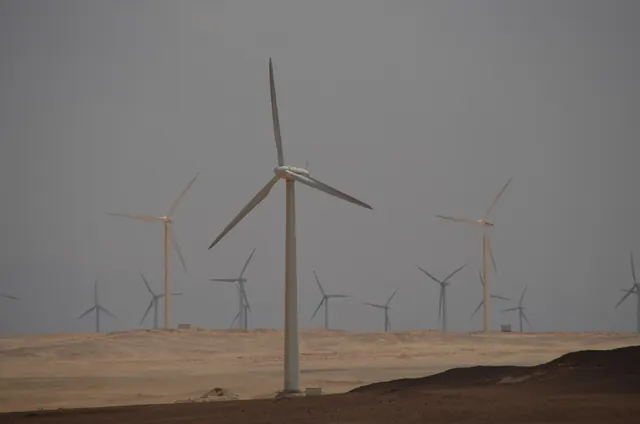 A wind turbine on sandy ground under a grey sky, with more turbines behind