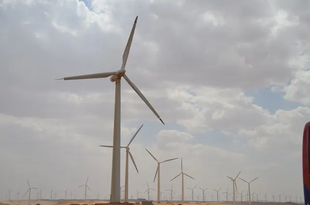 A tall wind turbine against white clouds, with rows of turbines on the horizon