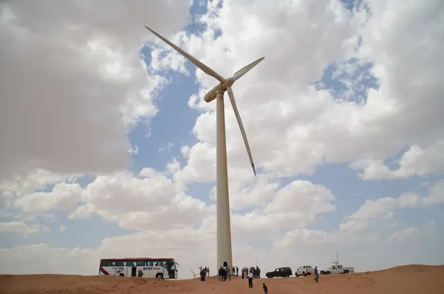 A wind turbine under a cloudy sky with a bus and visitors at its base