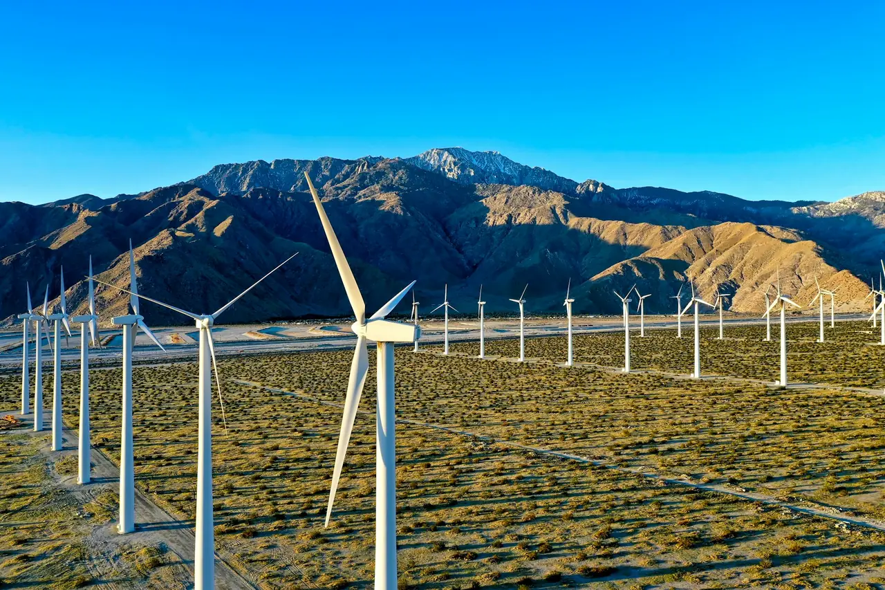 Rows of wind turbines on a scrub plain below bare mountains under a blue sky